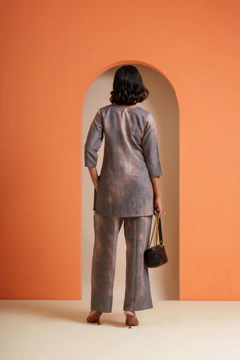 Woman in grey tie-dye co-ord set with flared pants, holding chain bag, back view against orange arch
